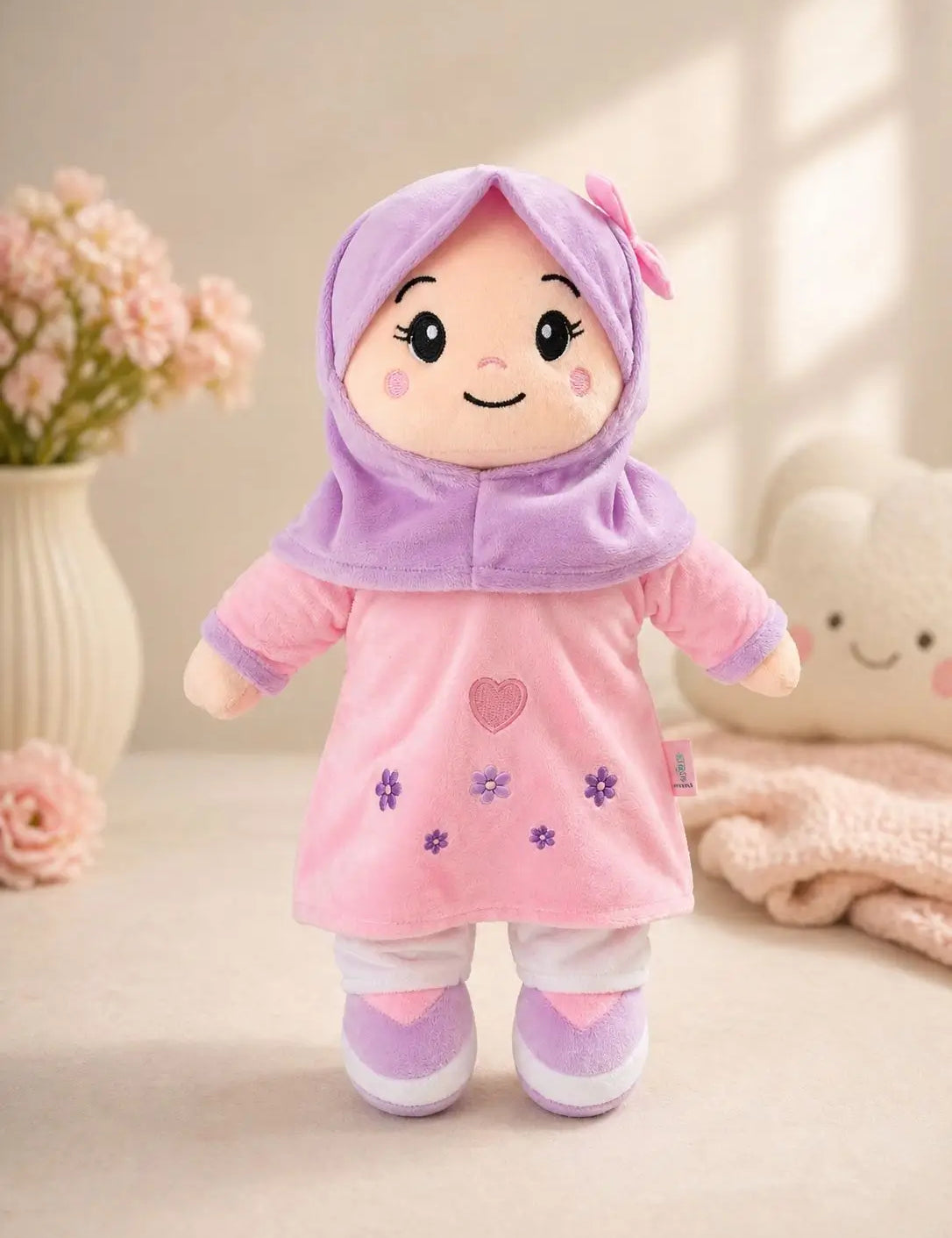 A plush doll wearing a pink dress and lavender-purple hijab, standing on a soft beige surface with decorative flowers and a plush toy in the background.