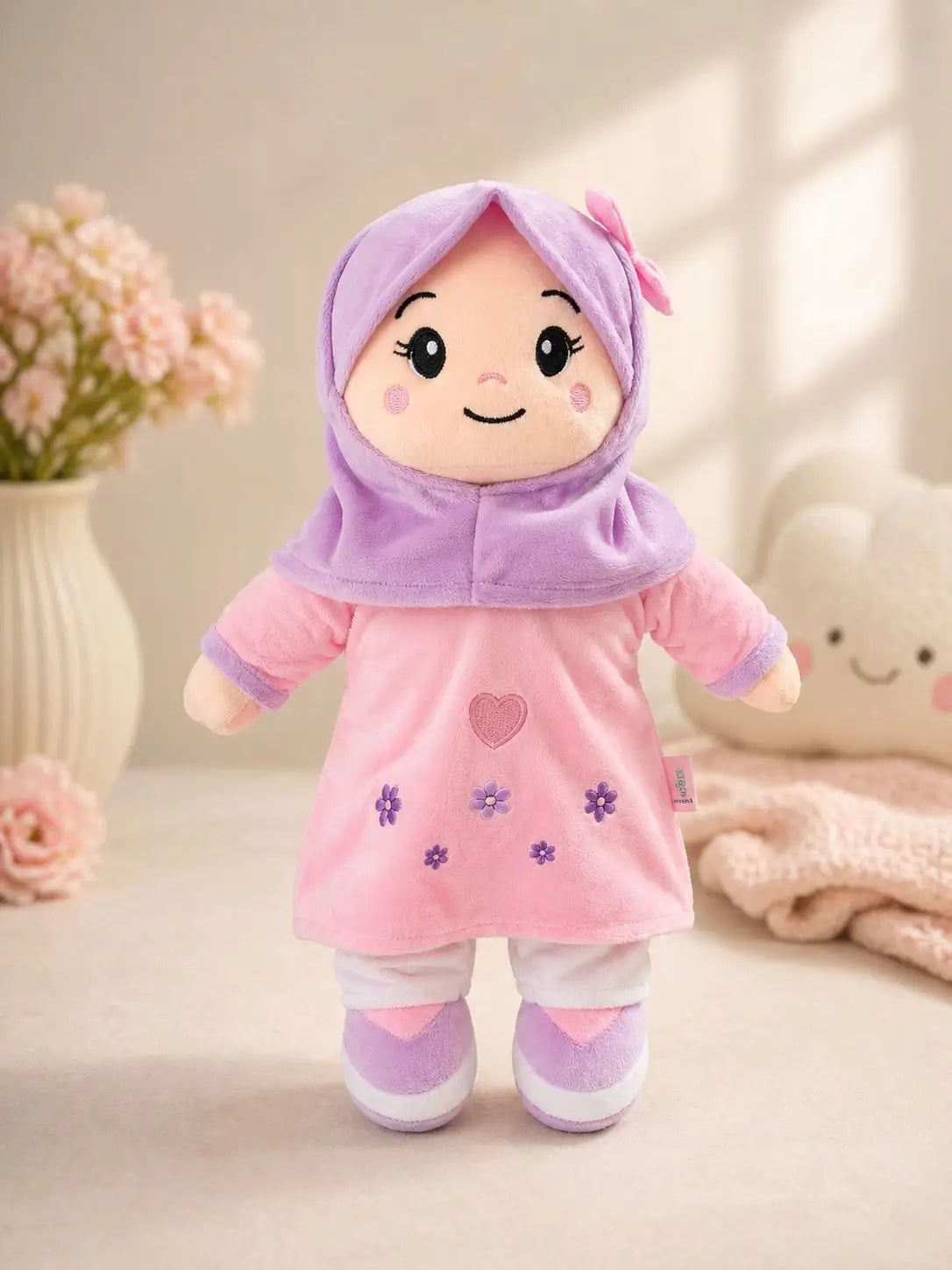 A plush doll wearing a pink dress and lavender-purple hijab, standing on a soft beige surface with decorative flowers and a plush toy in the background.