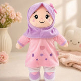 A plush doll wearing a pink dress and lavender-purple hijab, standing on a soft beige surface with decorative flowers and a plush toy in the background.
