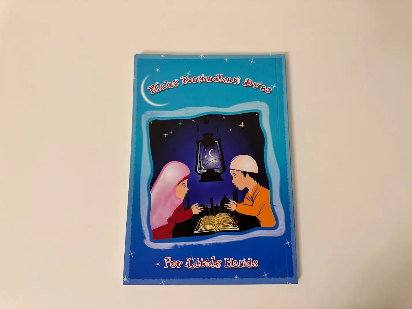 Children's book "Mahe Ramadhan Duas for Little Hands" with a blue cover featuring two children and a lamp on a beige background