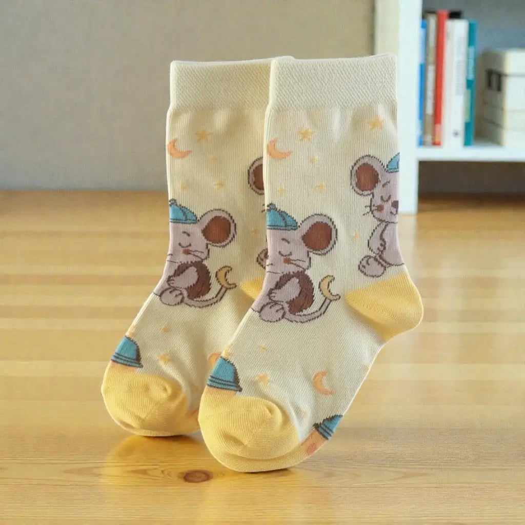 A pair of cream-colored baby socks with a cartoon mouse astronaut pattern, standing on a light wooden floor with a blurred bookshelf in the background.