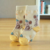 A pair of cream-colored baby socks with a cartoon mouse astronaut pattern, standing on a light wooden floor with a blurred bookshelf in the background.