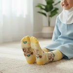 A smiling child wearing a light blue dress and white hijab sits on a cream rug indoors, with yellow socks featuring cartoon teddy bear prints prominently shown in the foreground.