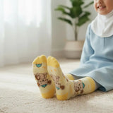A smiling child wearing a light blue dress and white hijab sits on a cream rug indoors, with yellow socks featuring cartoon teddy bear prints prominently shown in the foreground.