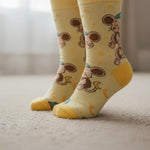 A pair of pale yellow baby socks with a cartoon monkey and duck pattern, shown on a child's feet standing on a light-colored carpet.