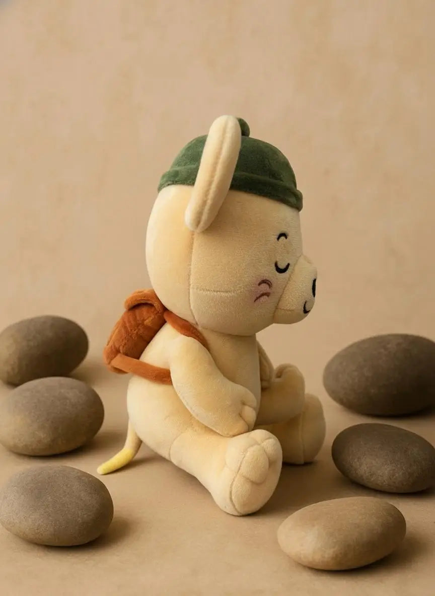 A plush toy character sitting among smooth stones on a beige surface, wearing a dark green hat and orange backpack.