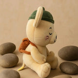 A plush toy character sitting among smooth stones on a beige surface, wearing a dark green hat and orange backpack.