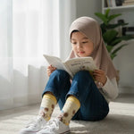 A young girl wearing a beige hijab, blue jeans, and white sneakers sits on a cream rug, reading a book in a bright, sunlit room.