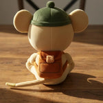 A plush mouse toy shown from the back, wearing a green cap and orange backpack, sitting on a wooden table in warm sunlight.