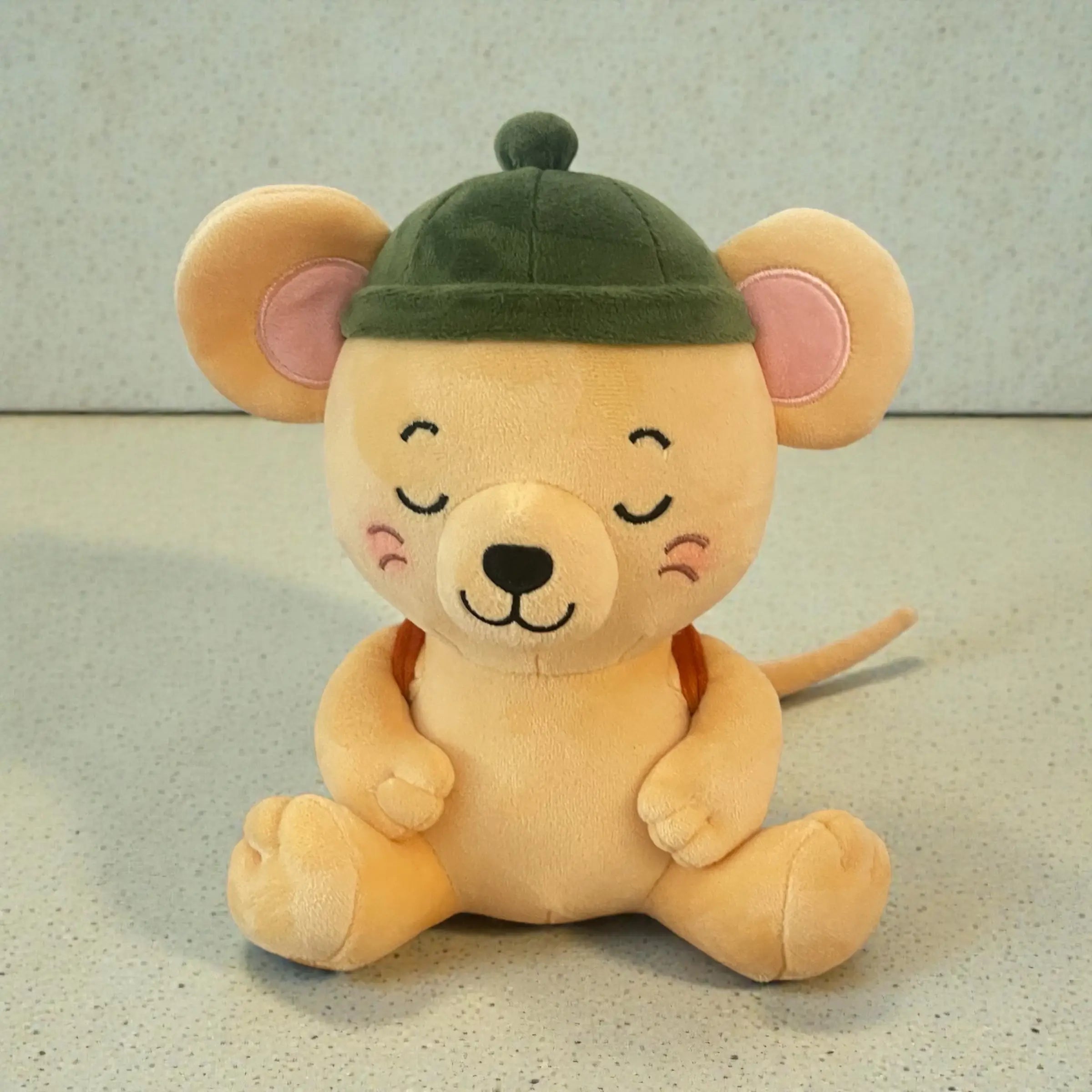 A plush toy mouse sitting down, wearing a dark green cap. The toy has a warm beige body, pink inner ears, and embroidered closed eyes and smiling mouth.
