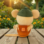 A plush mouse toy shown from the back, sitting on a wooden table outdoors with sunflowers and greenery in the background. The plush has a cream-colored body, large ears, a dark green cap, and an orange backpack.