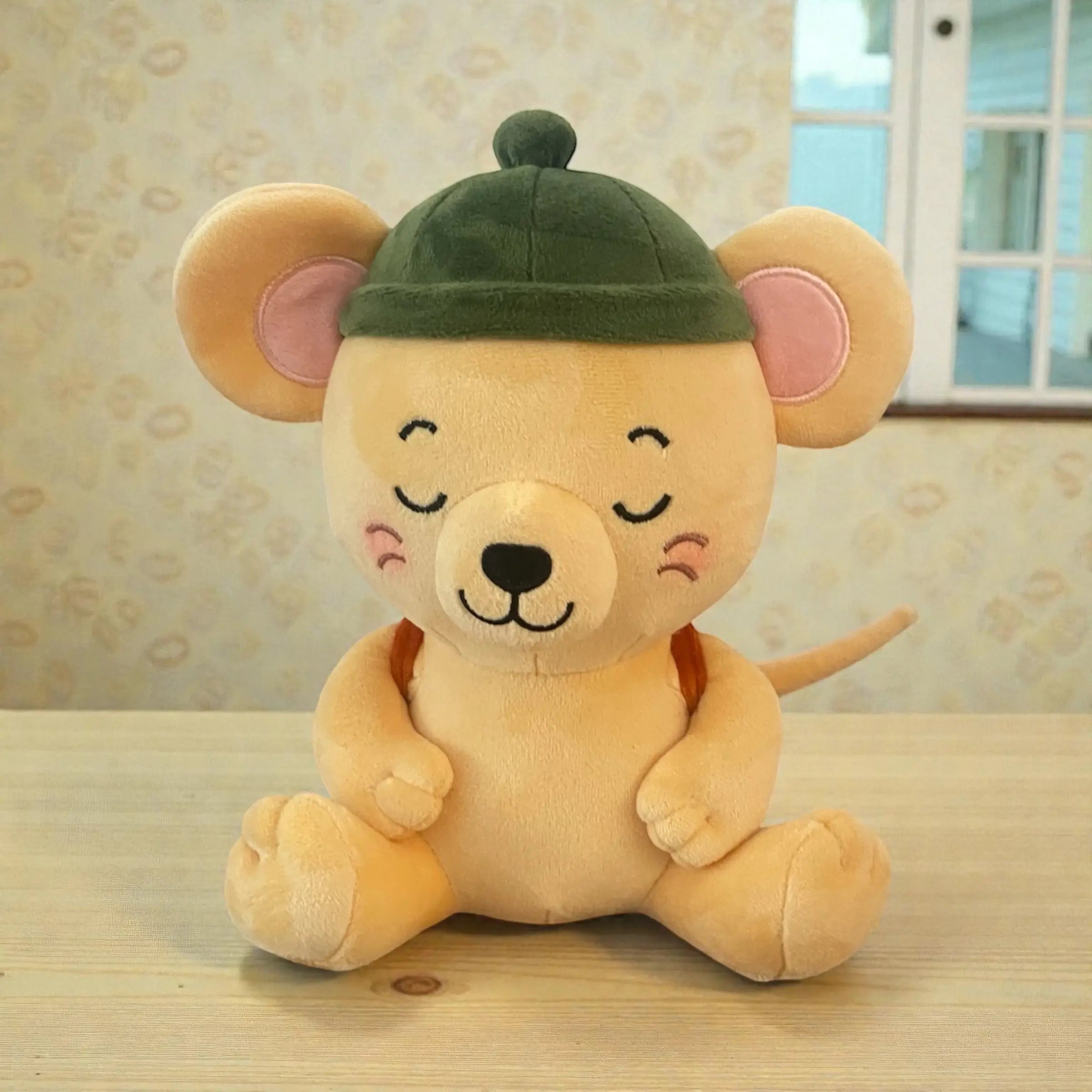 A plush toy mouse sitting on a light wooden table, wearing a dark green beret-style hat. The toy has a warm beige body, pink inner ears, closed embroidered eyes, and a small orange-brown backpack, set against a patterned wallpaper background.