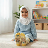 A young child wearing a light blue modest outfit, white hijab, and Aliya and Hasan Bookshelf yellow socks sits smiling on a cream rug in a bright, modern playroom.