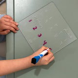 A child's hands holding a transparent magnetic numbers board with Arabic numerals and writing on it with a blue and white marker.