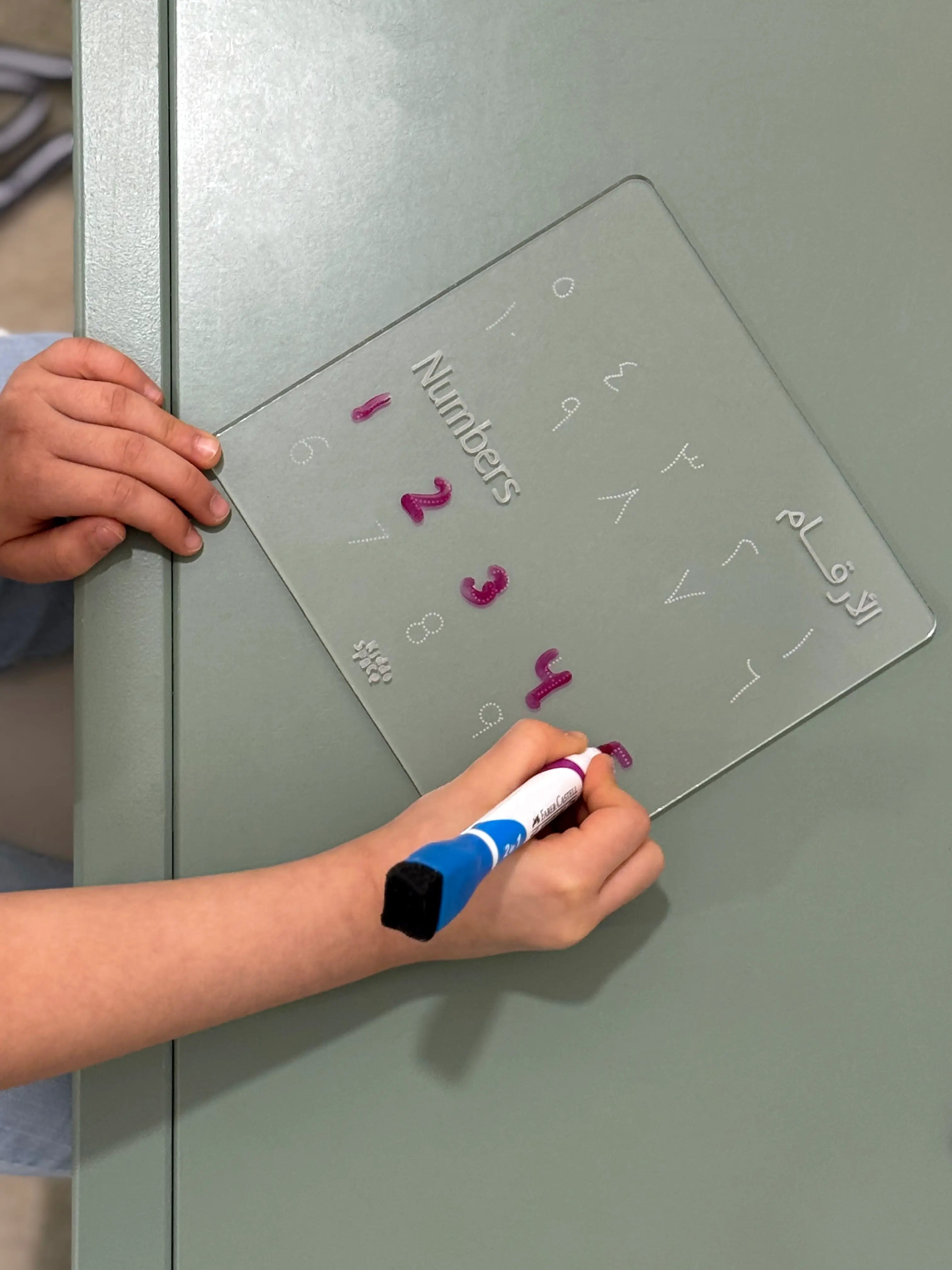 A child's hands holding a transparent magnetic numbers board with Arabic numerals and writing on it with a blue and white marker.