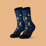 A pair of dark navy blue crew socks with a festive Ramadan pattern featuring yellow crescent moons, stars, lanterns, and mosque silhouettes, displayed against a solid peach background.
