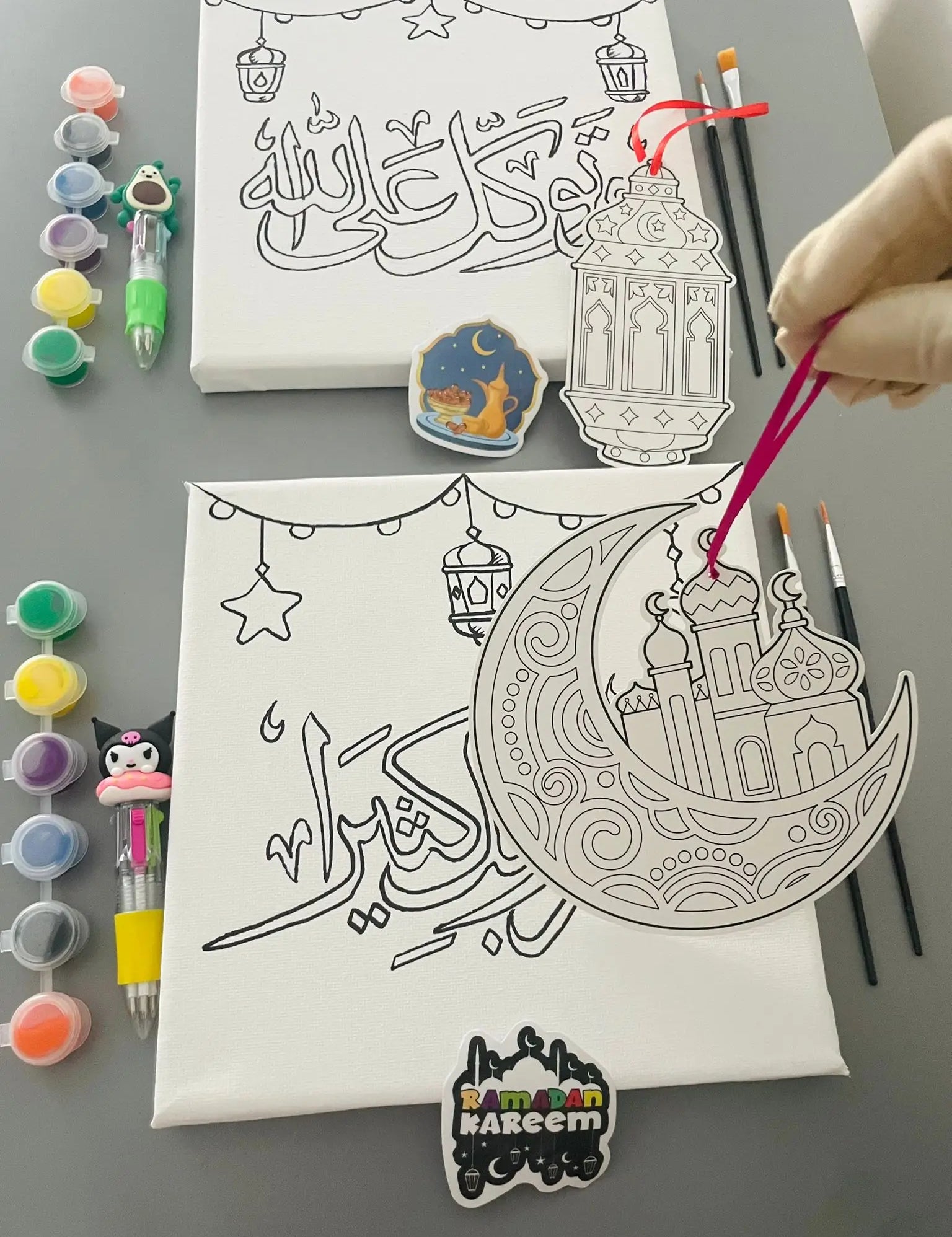 Canvas with crescent moon and star design, paint bottles, and brushes on a gray surface.