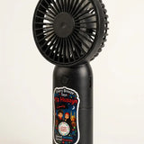 A black handheld personal fan with a circular fan head and a cylindrical body, featuring a colorful printed label on the front, photographed against a plain white background.
