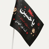 A small black flag on a white stick, featuring Arabic text in white and red with cartoon children and red hearts, shown against a light gray background.