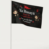 A small black flag on a white pole with a gold finial, printed with cartoon child figures, red hearts, and the text 'Ya Husayn Peace be upon you You are the light of guidance' on a light gray background.