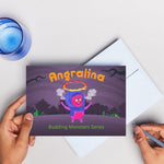 Book with 'Angralina' design held by hands on a light surface with a mug and stickers.