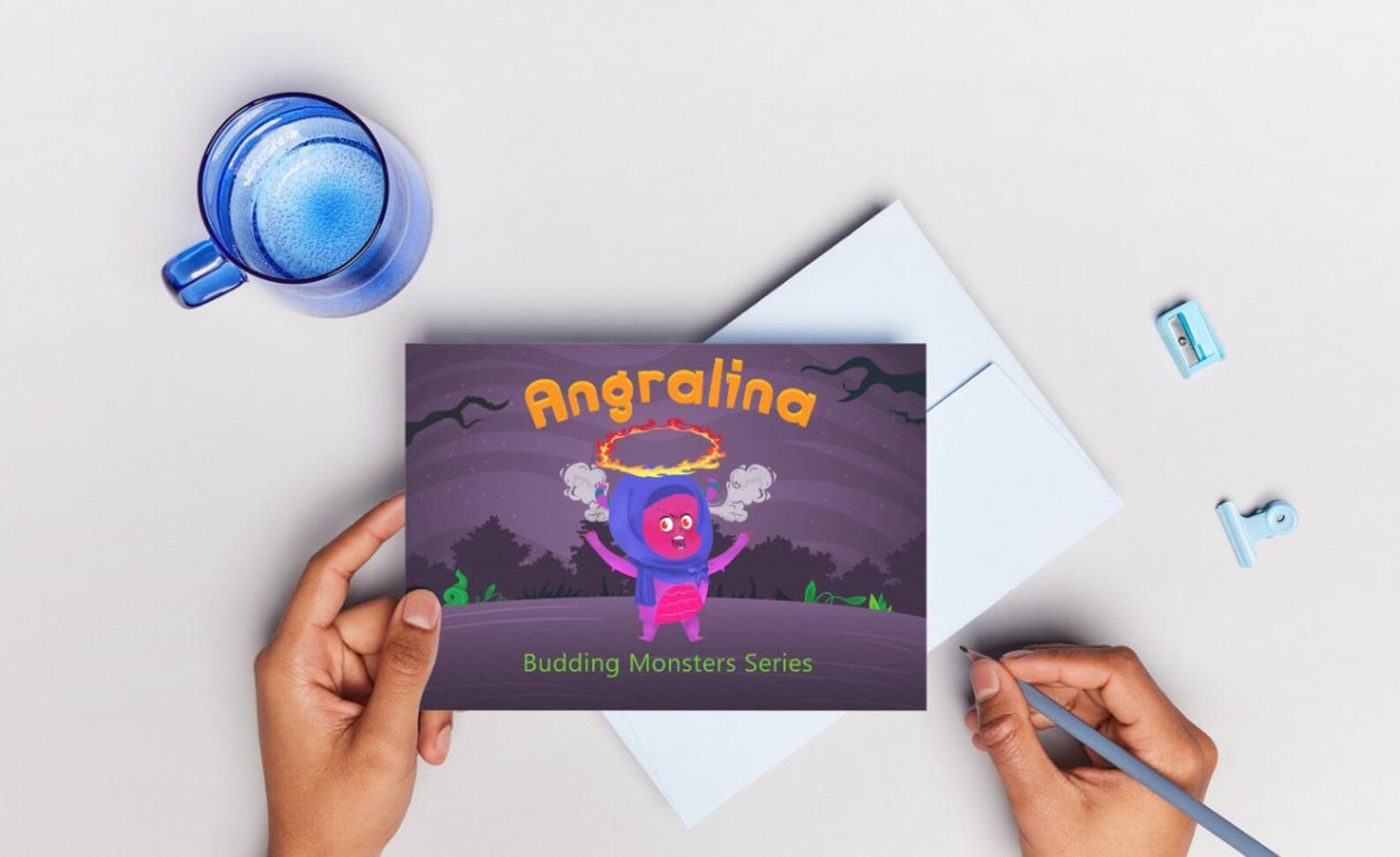 Book with 'Angralina' design held by hands on a light surface with a mug and stickers.