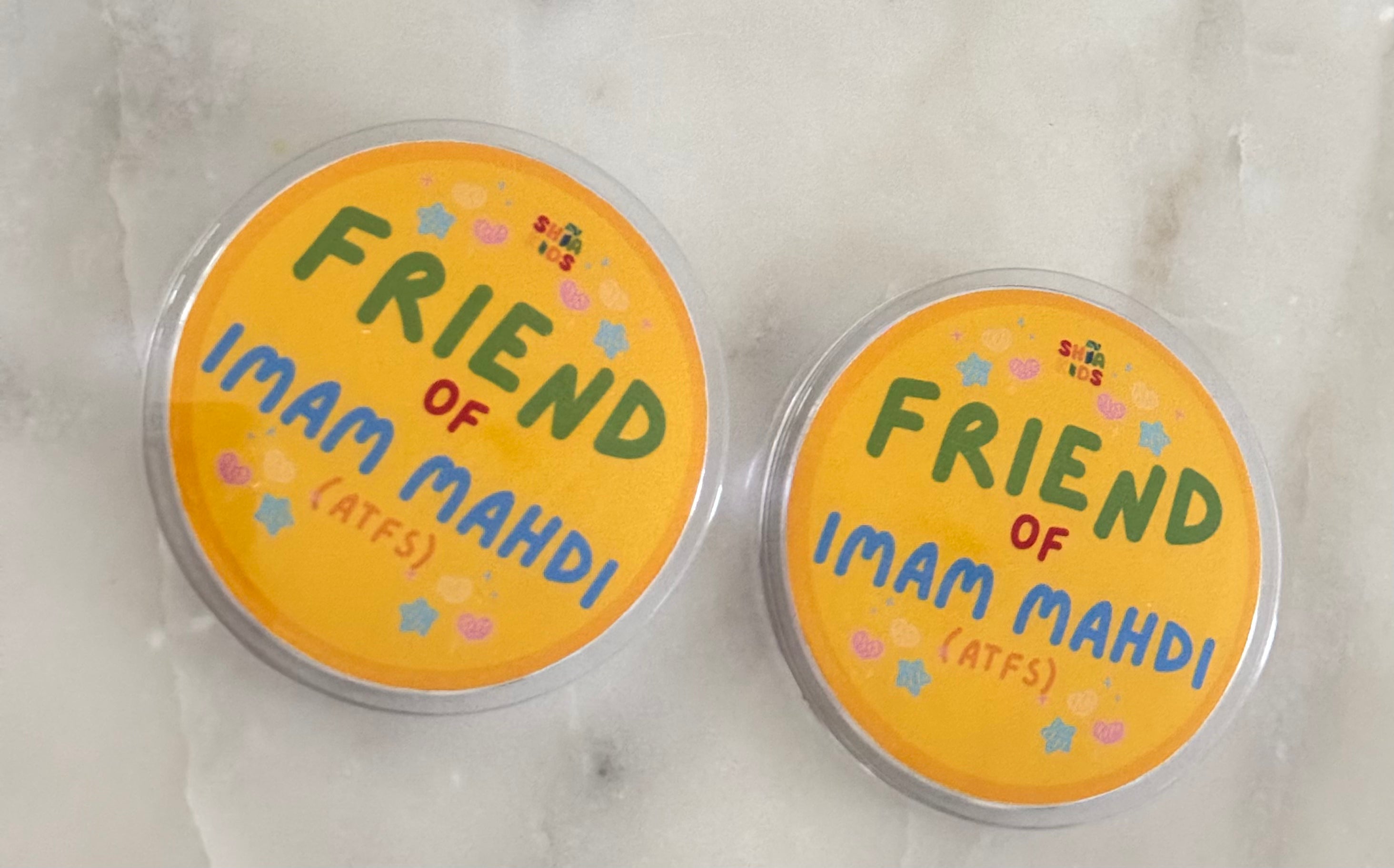 Two round badges with the text 'FRIEND OF IMAM MAHDI (ATFS)' written on them, placed on a marble surface.