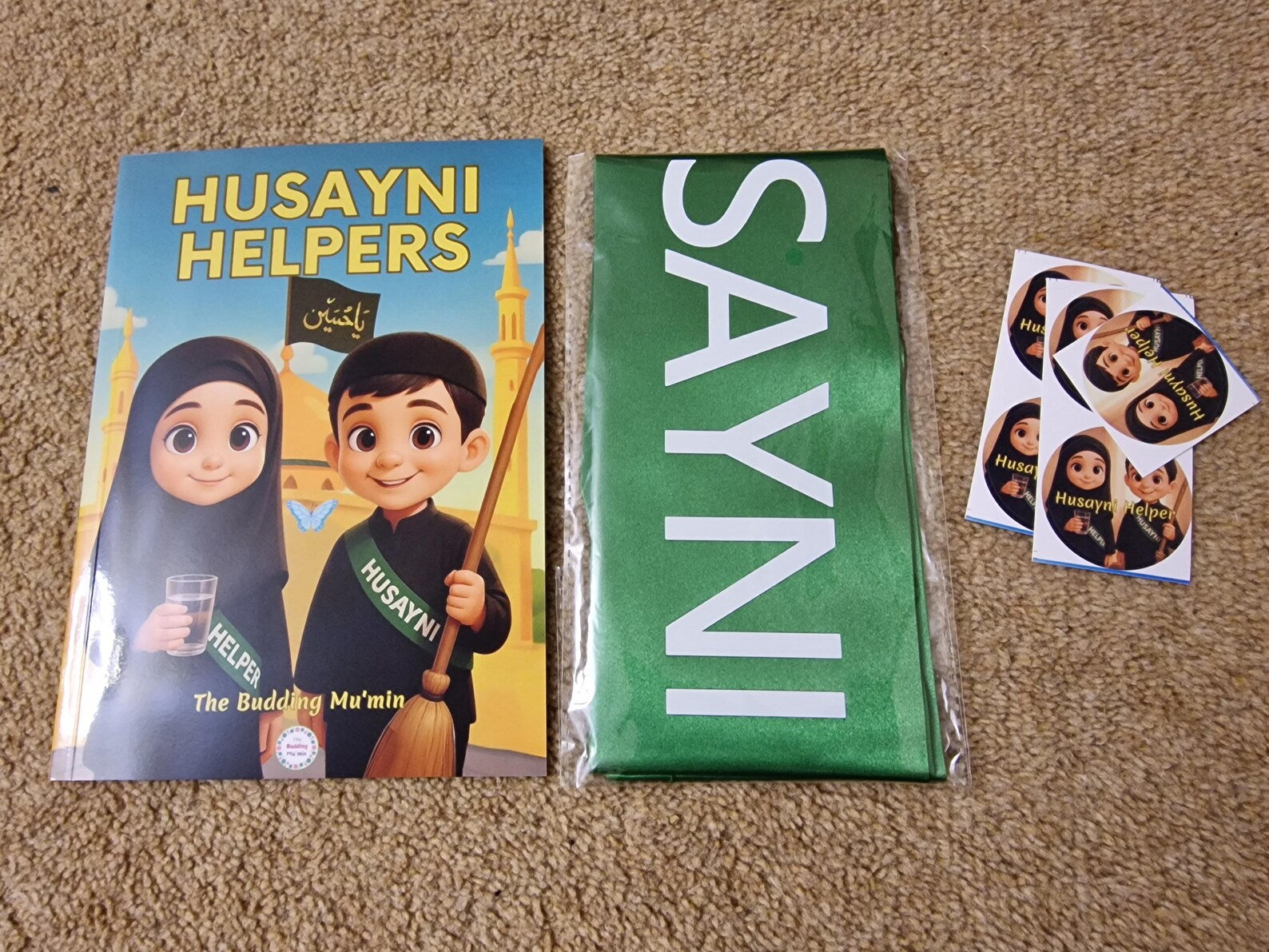 Book titled 'Husayni Helpers' with a green sash and stickers on a brown carpet.