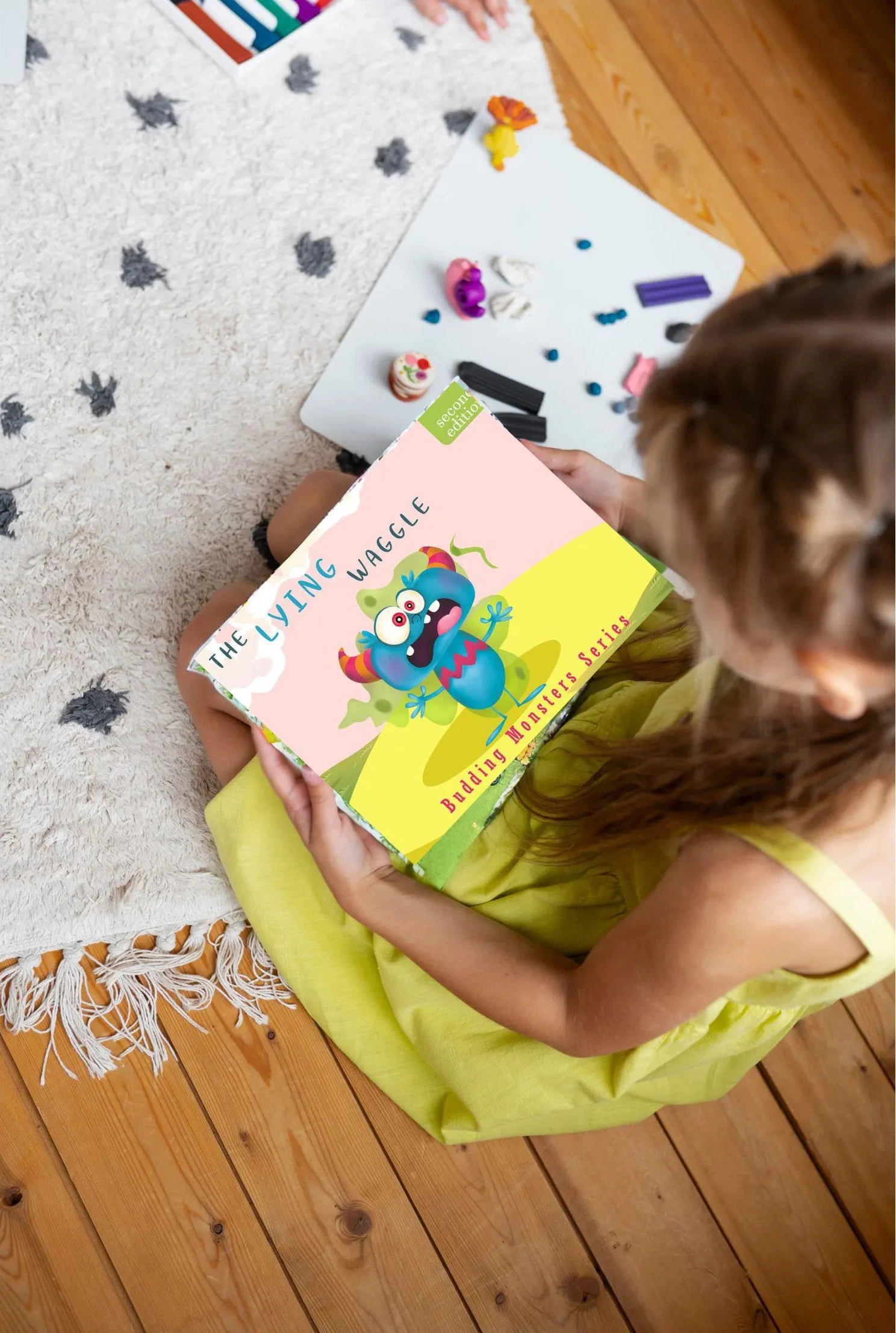 Child holding a colourful book from the Budding Monsters series on a wooden floor with toys scattered around